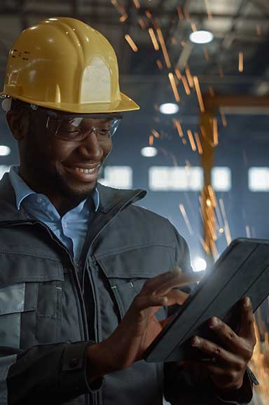Homem negro com capacete industrial amarelo, sorrindo e mexendo no tablet
